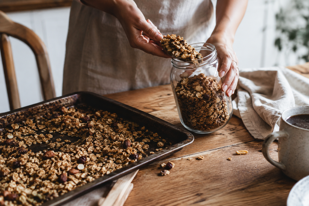 Hand pouring freshly baked homemade granola into a glass jar on a lived-in kitchen table.