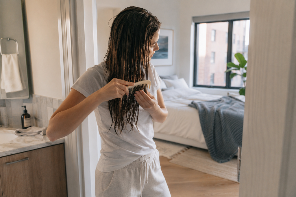 Woman gently combing damp hair after a Sunday evening shower in a bright apartment bedroom.