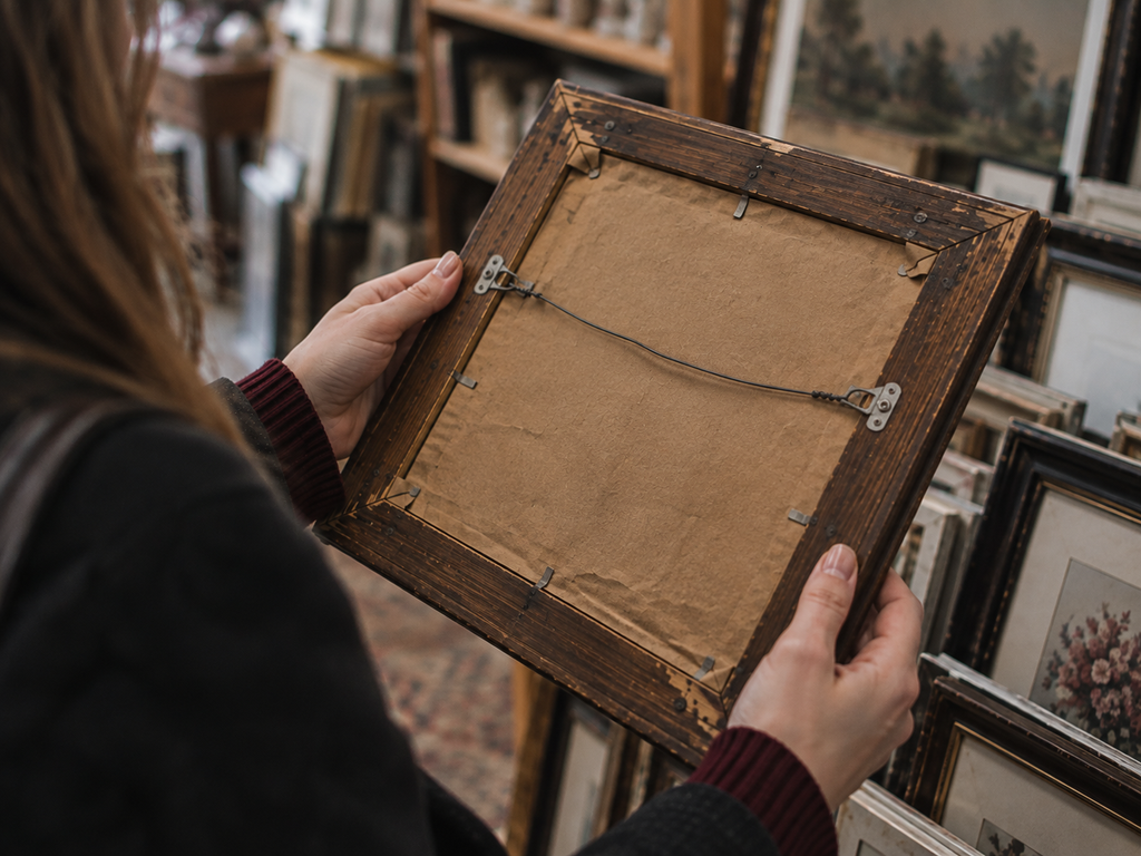 Hands inspecting the back, corner joints and hanging wire of an old thrift-store picture frame.